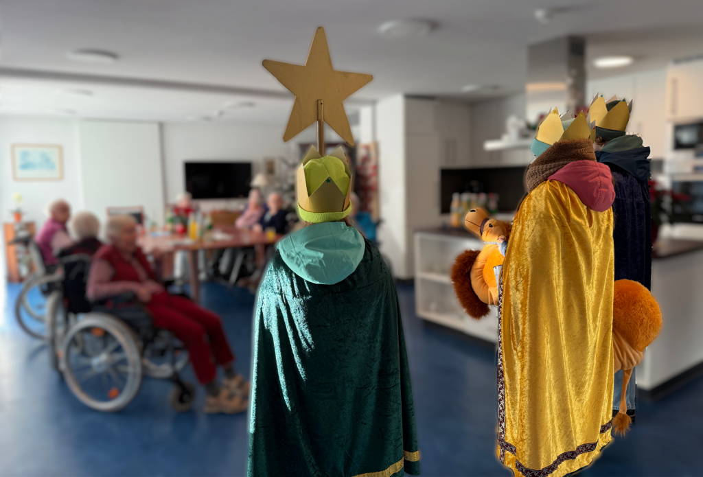 Drei Jugendliche, als Sternsinger verkleidet, stehen mit dem Rücken zur Kamera im Gruppenraum der Pflege-Einrichtung. Im Hintergrund sind unscharf sitzende Menschen zu erkennen, die den Sternsingern zugewandt sind.