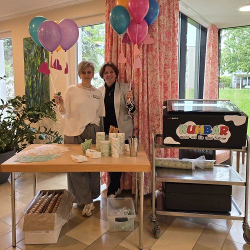 Zwei lächelnde Frauen stehen vor einem mit Eis und Grußkarten gedeckten Tisch und halten Luftballons in den Händen.