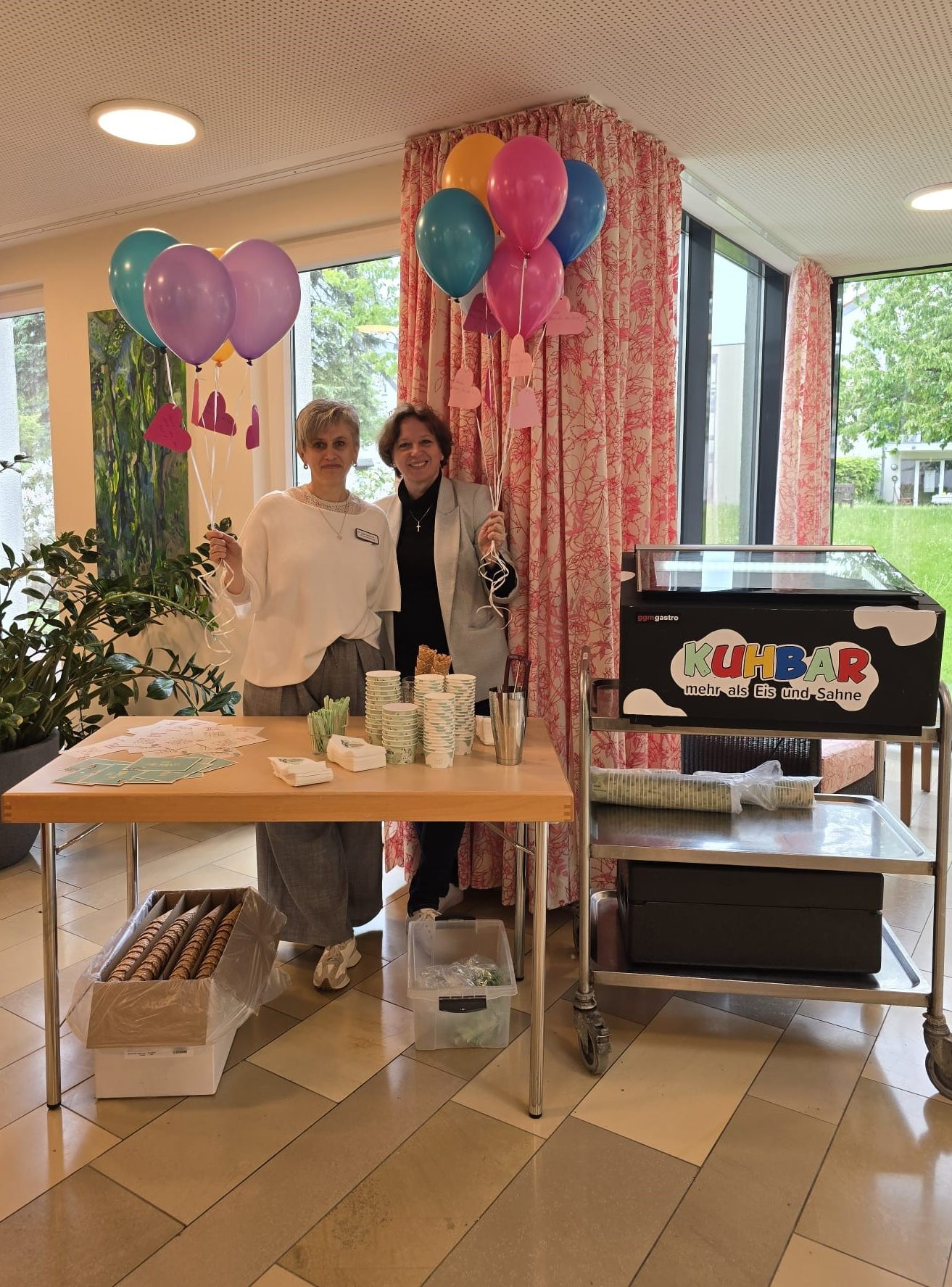 Zwei lächelnde Frauen stehen vor einem mit Eis und Grußkarten gedeckten Tisch und halten Luftballons in den Händen.